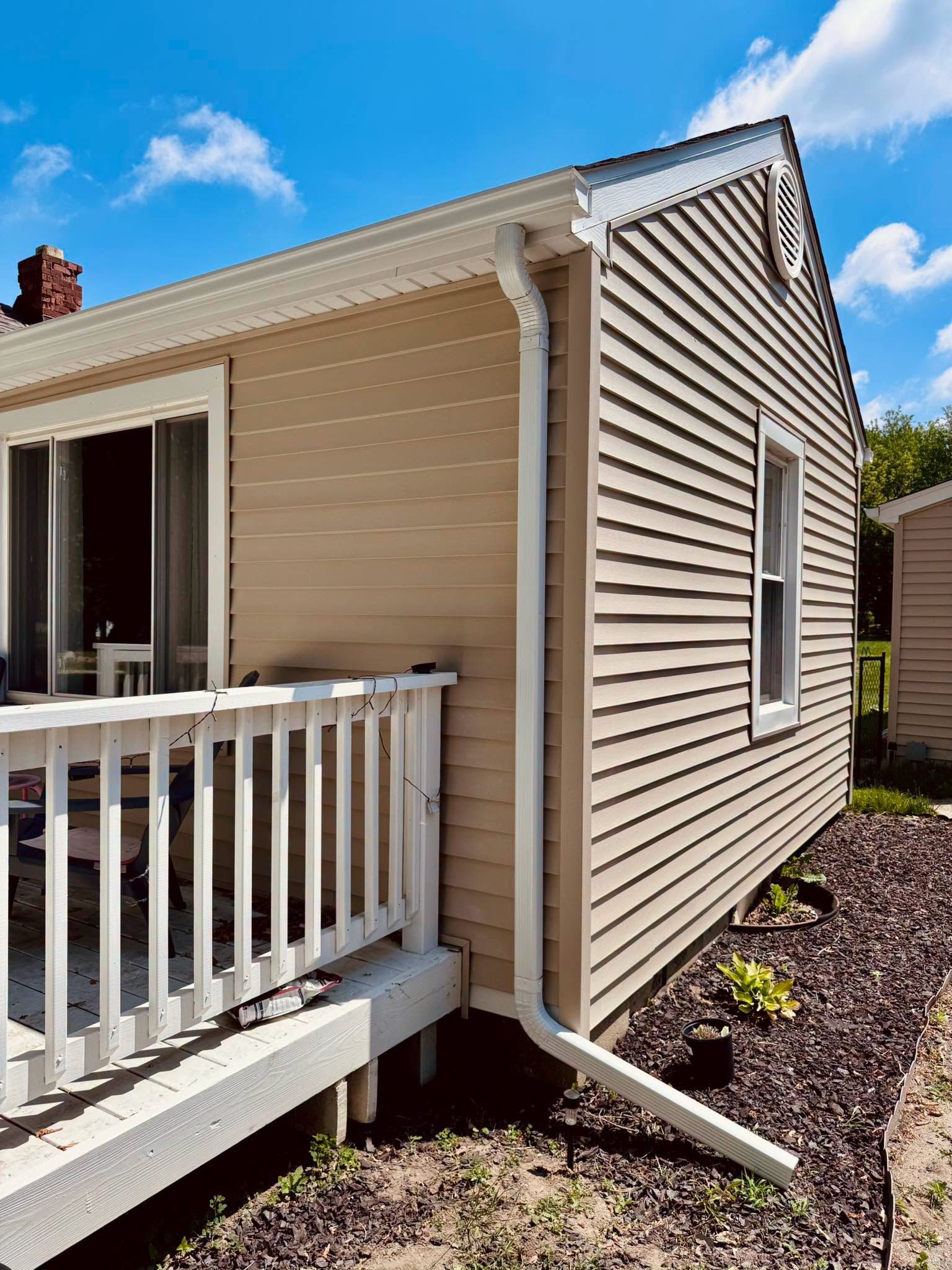 Exterior view of a beige house with white porch railing, siding, and downspout beside a landscaped yard.