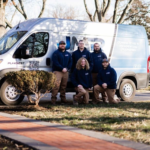 Five people in navy uniforms posing in front of a white work van outdoors on a sunny day.