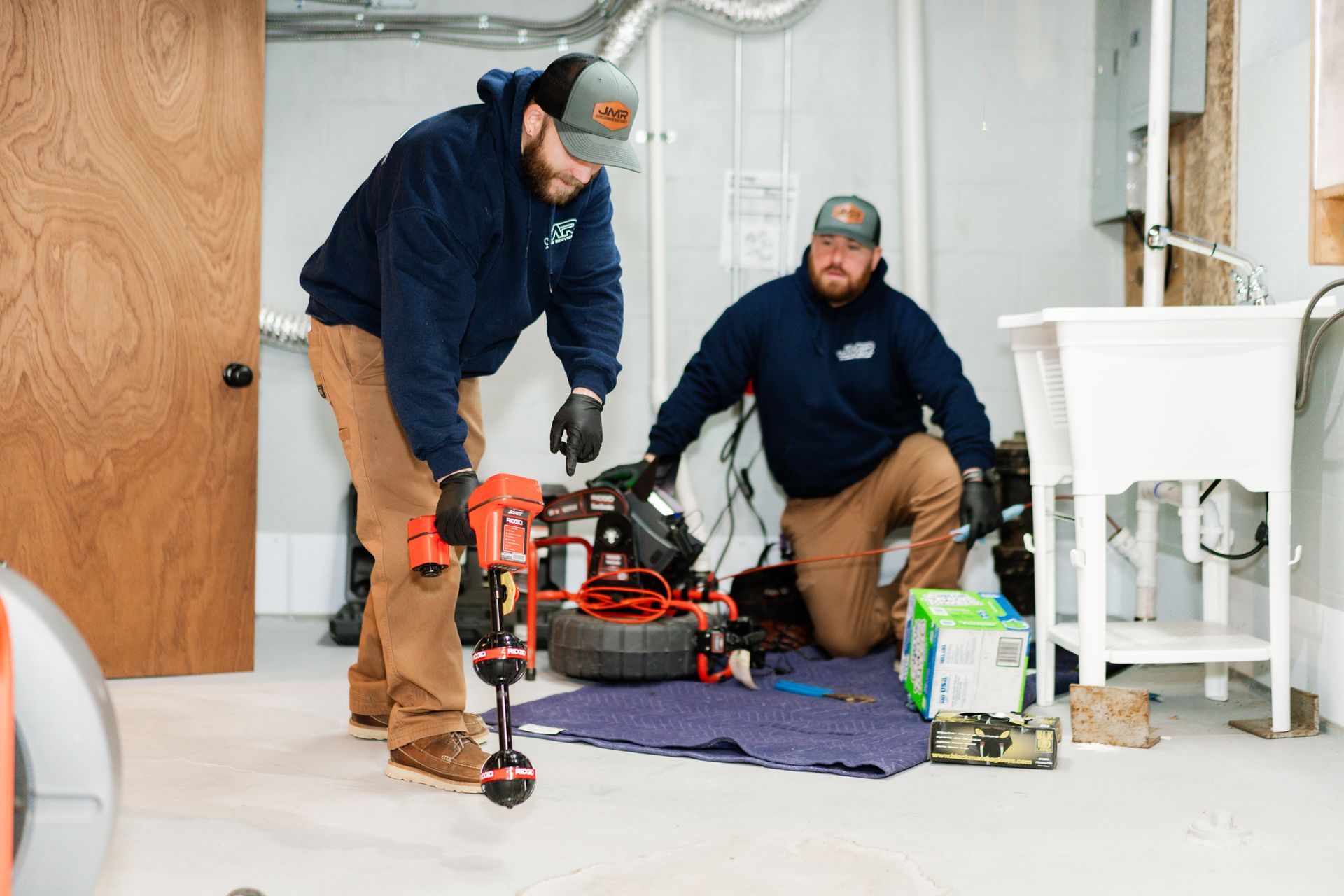 Two service technicians in blue uniforms operate a drain cleaning machine in an indoor utility room.