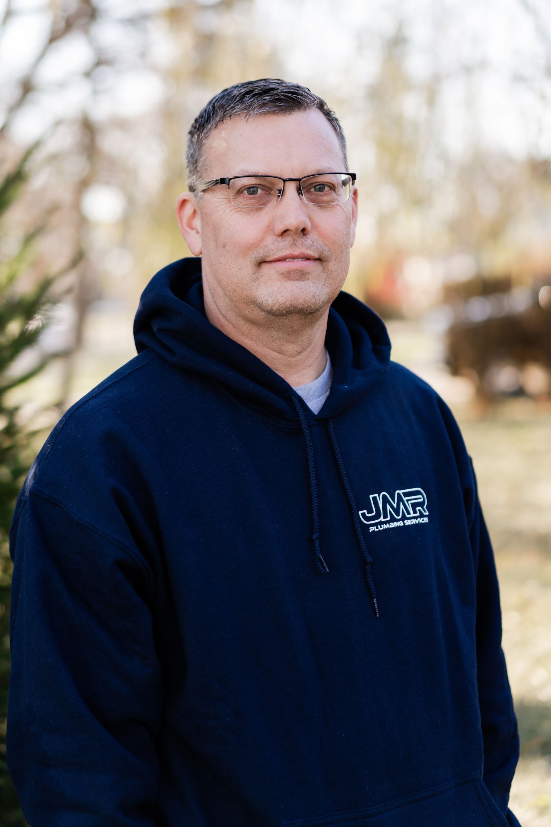 A person wearing glasses and a navy blue hoodie stands outdoors in a soft-focus, natural setting.