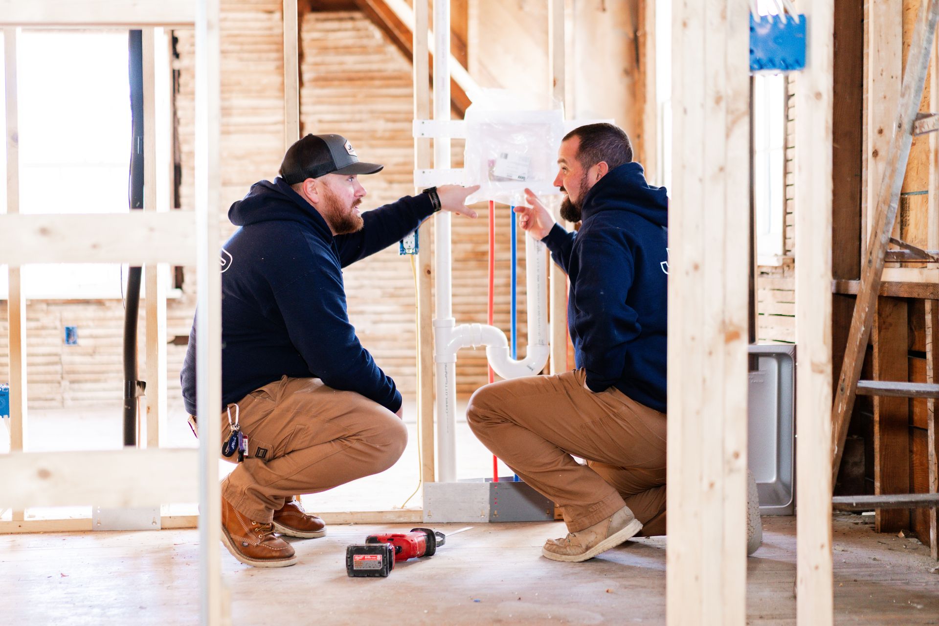 Two construction workers in dark hoodies and tan pants crouch in a wooden-framed house, discussing plumbing installation.