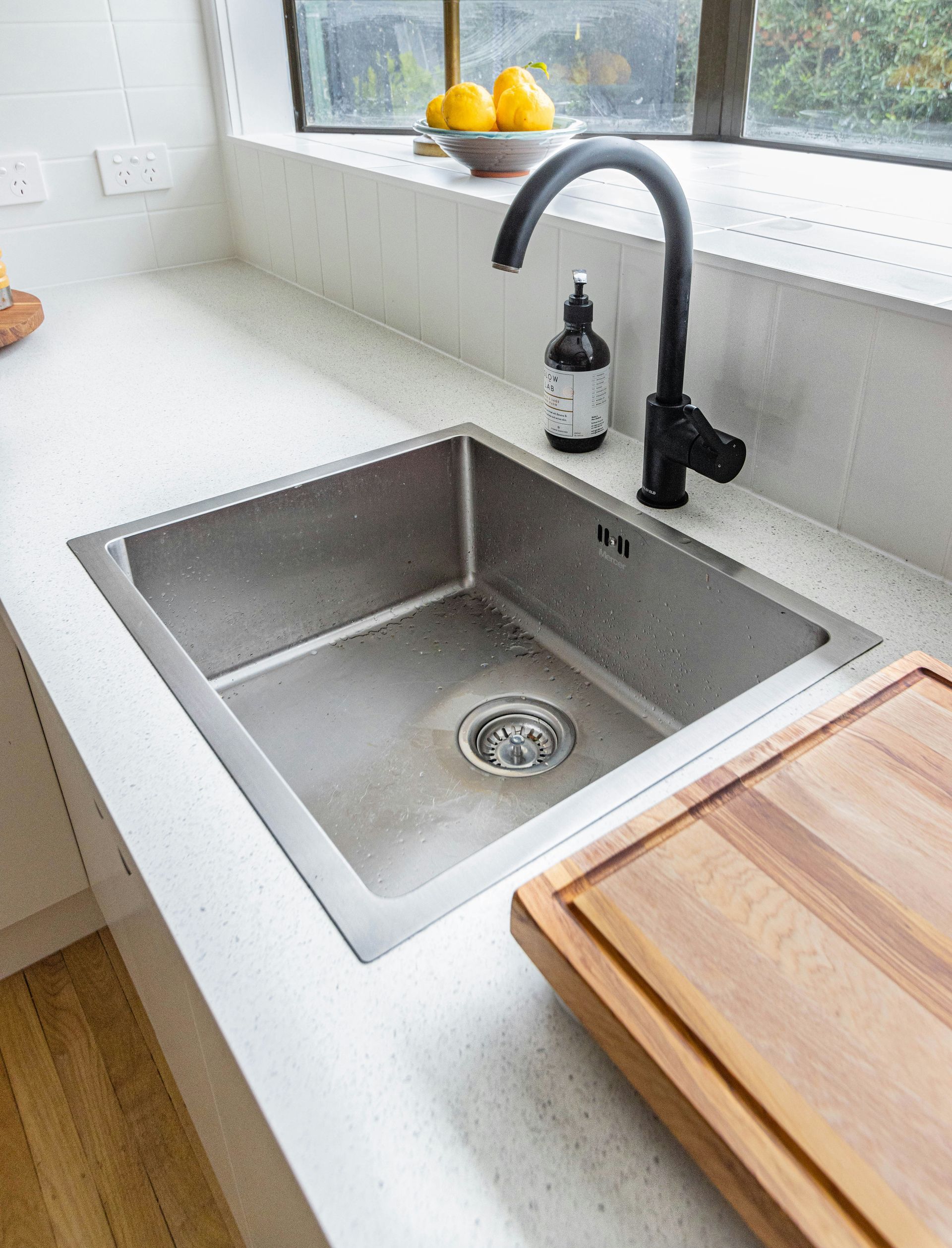 A stainless steel kitchen sink set into a white countertop, featuring a black gooseneck faucet and a wooden cutting board.