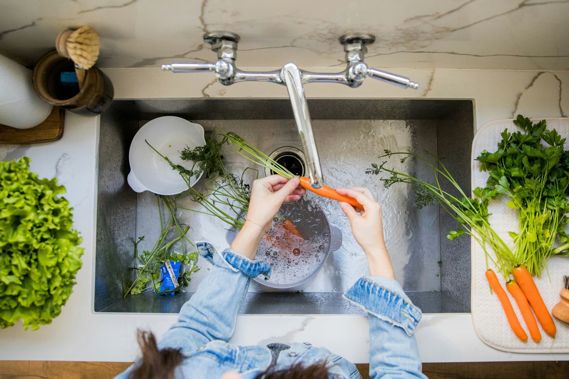 A person in a denim jacket rinses a carrot under the faucet in a kitchen sink next to fresh produce.