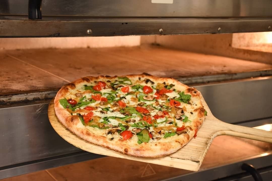Pizza on a wooden peel inside a brick oven, topped with cheese, tomatoes, and herbs.