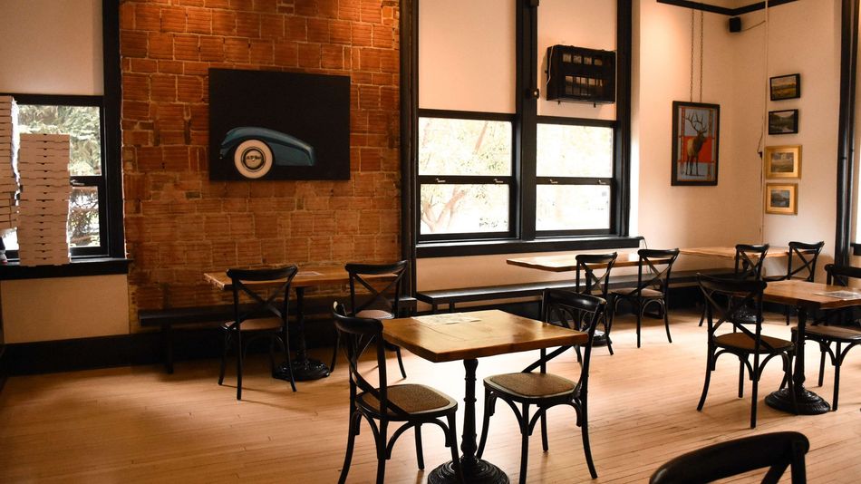 Empty café seating area with black chairs, wooden tables, brick walls, and large windows.