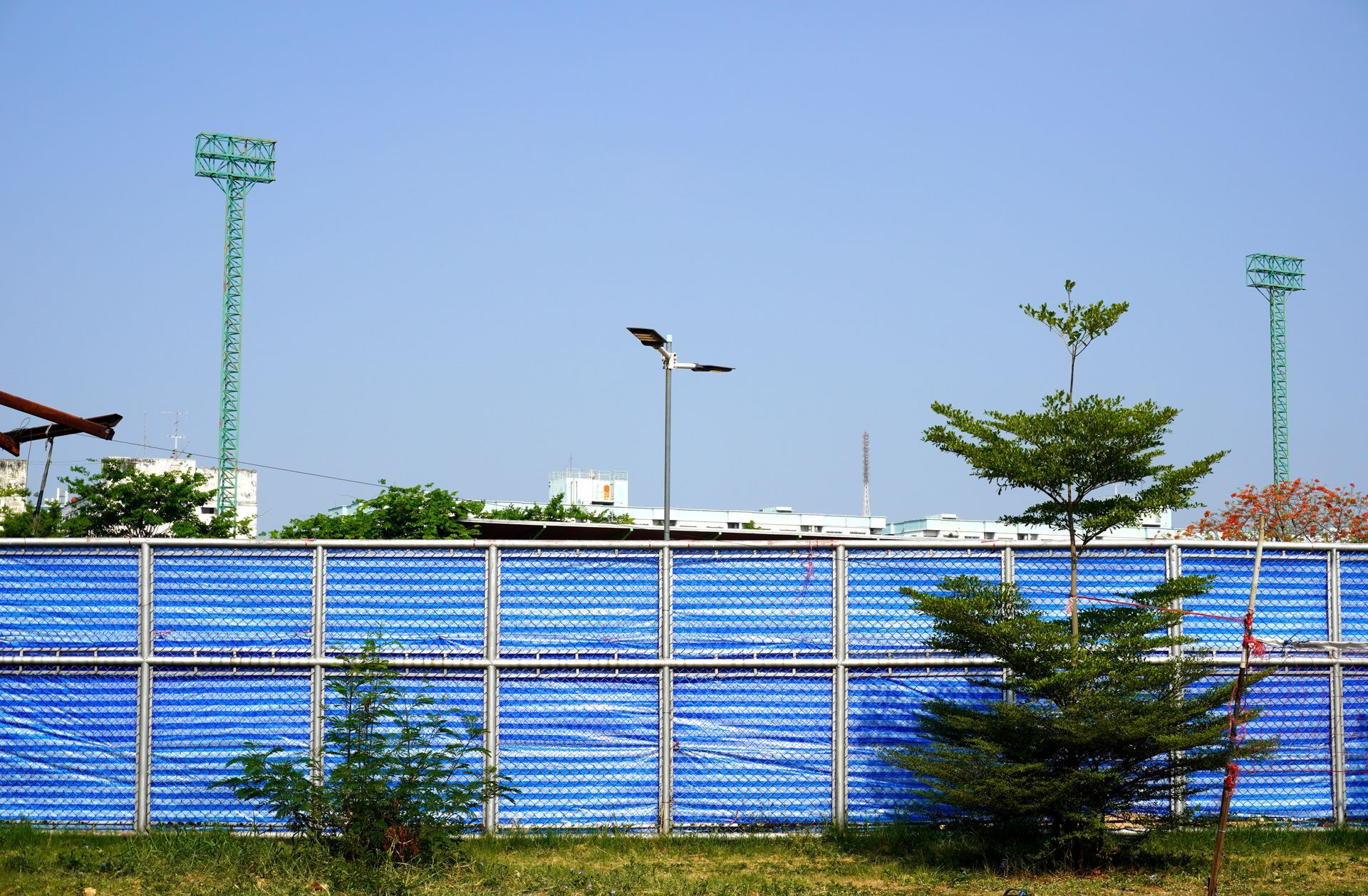 Blue fence in front of a field with trees, tall stadium lights, and a clear sky