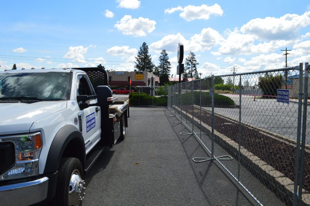 Open gate in a wire fence beside a dirt path and driveway, with trees and shrubs beyond.