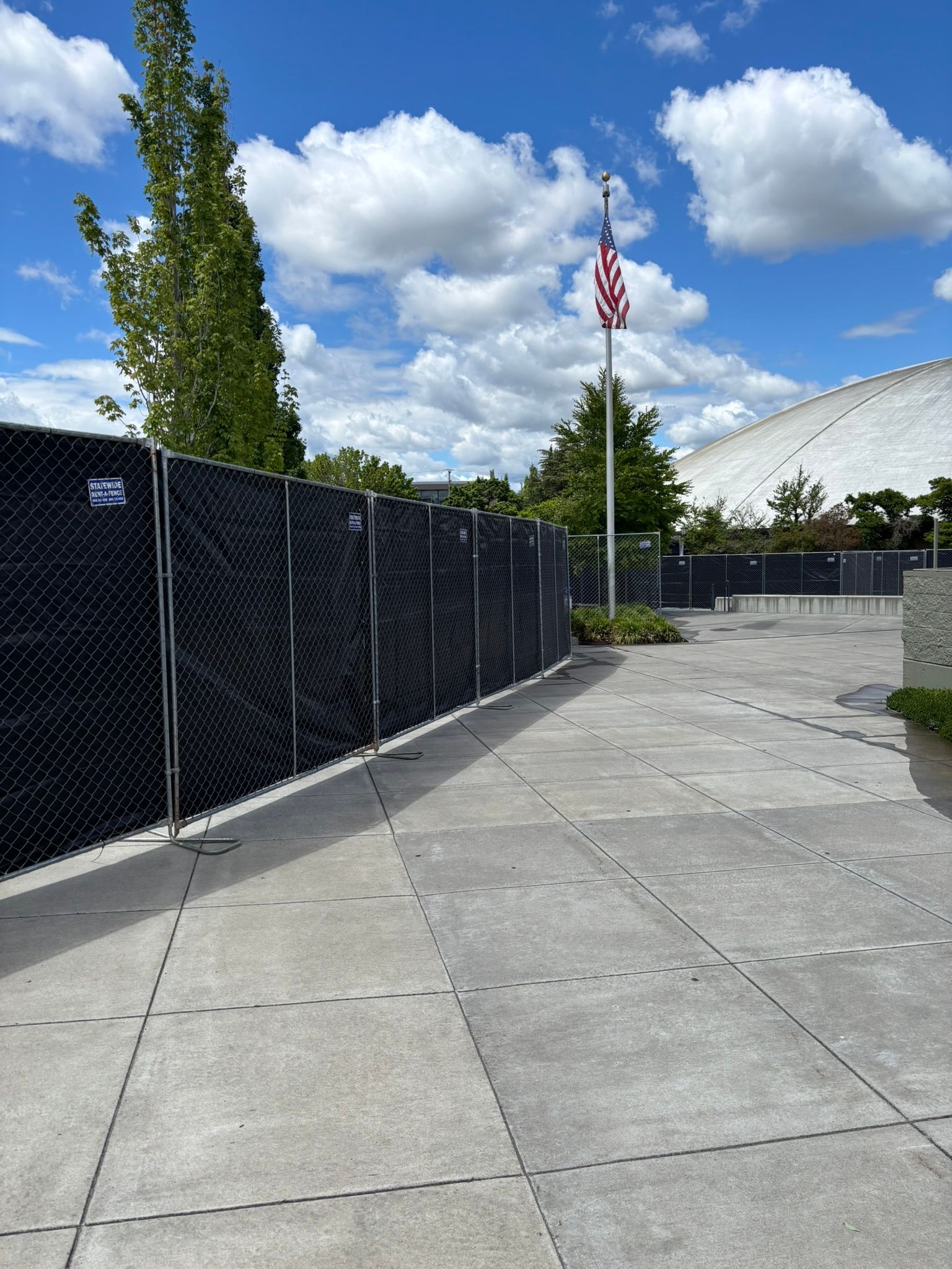 Empty paved walkway beside a black fence and U.S. flag under a blue sky with clouds