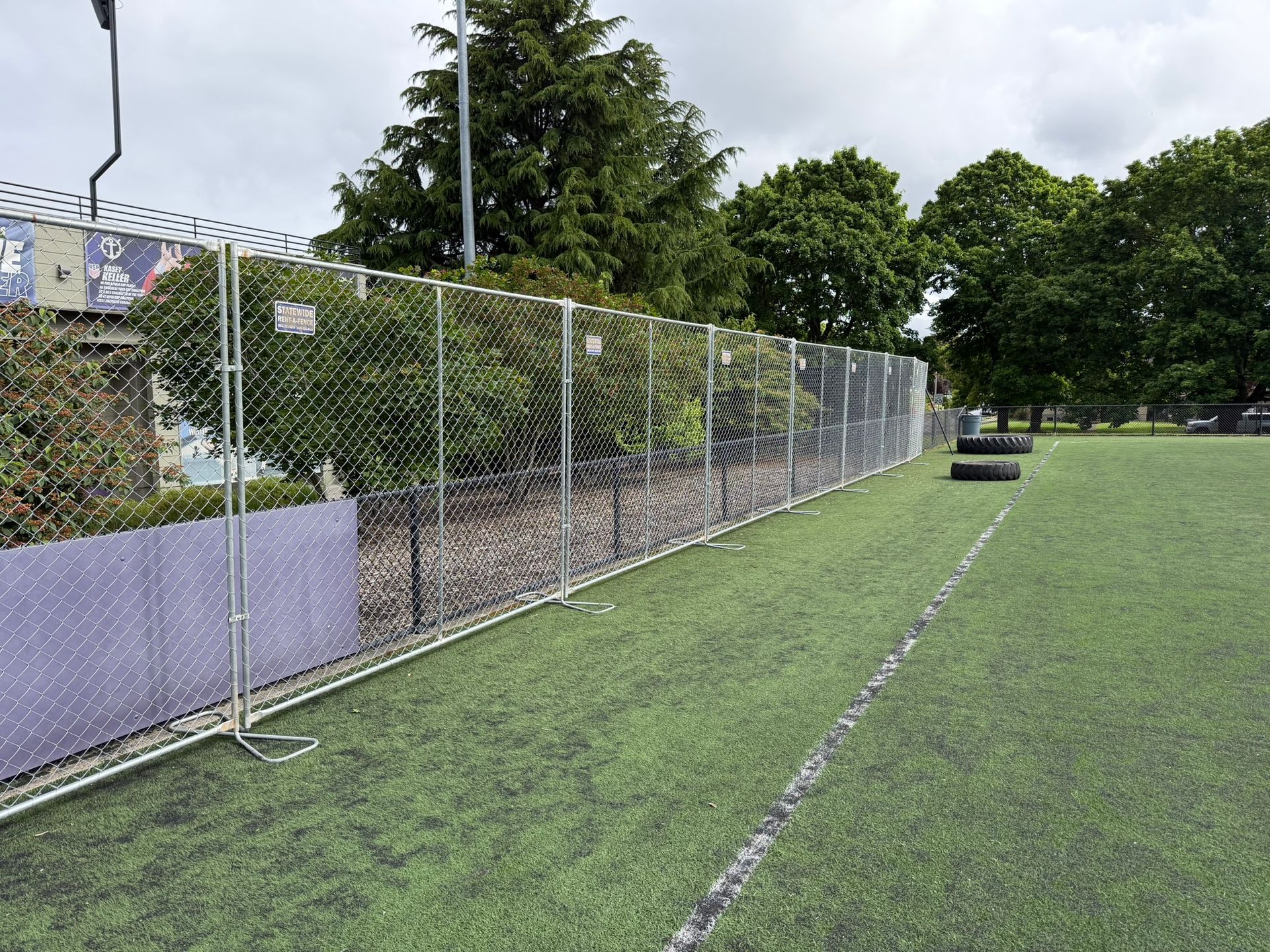Empty fenced area beside a paved road with trees and hedges under a blue sky