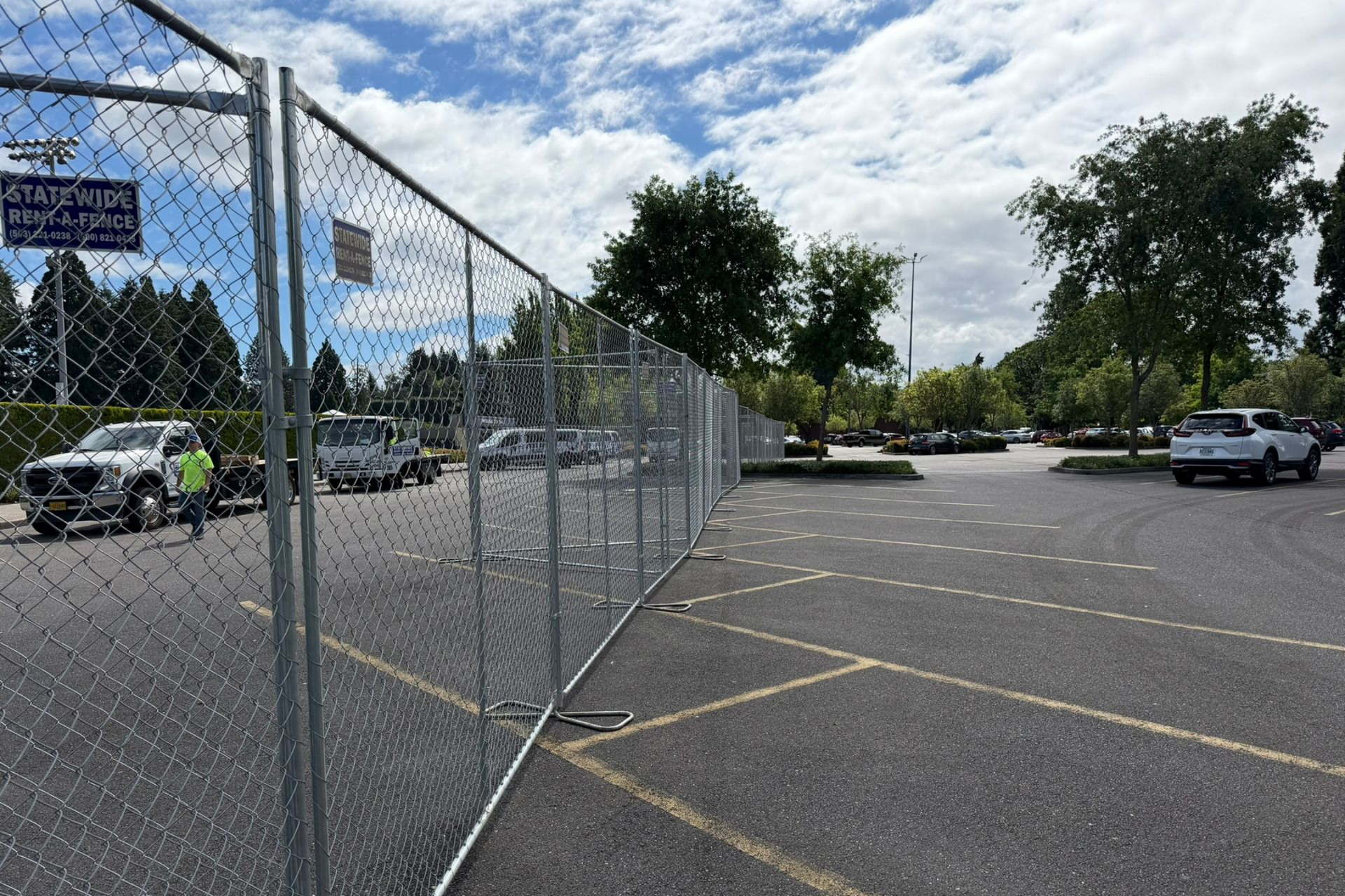 White crowd-control barriers along a wet sidewalk beside a grassy park, with trees in the background
