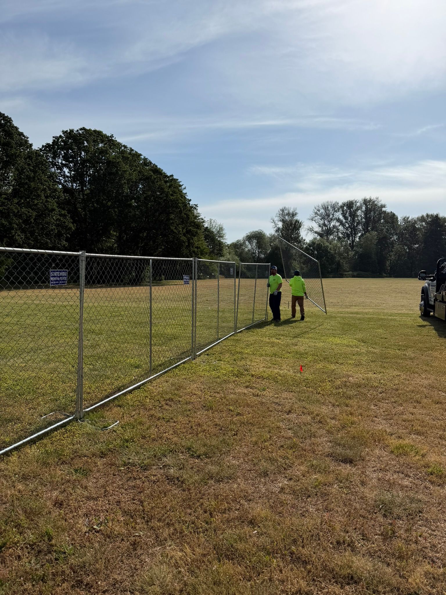 Three people in neon vests stand by a chain-link fence on a sunny grassy field.