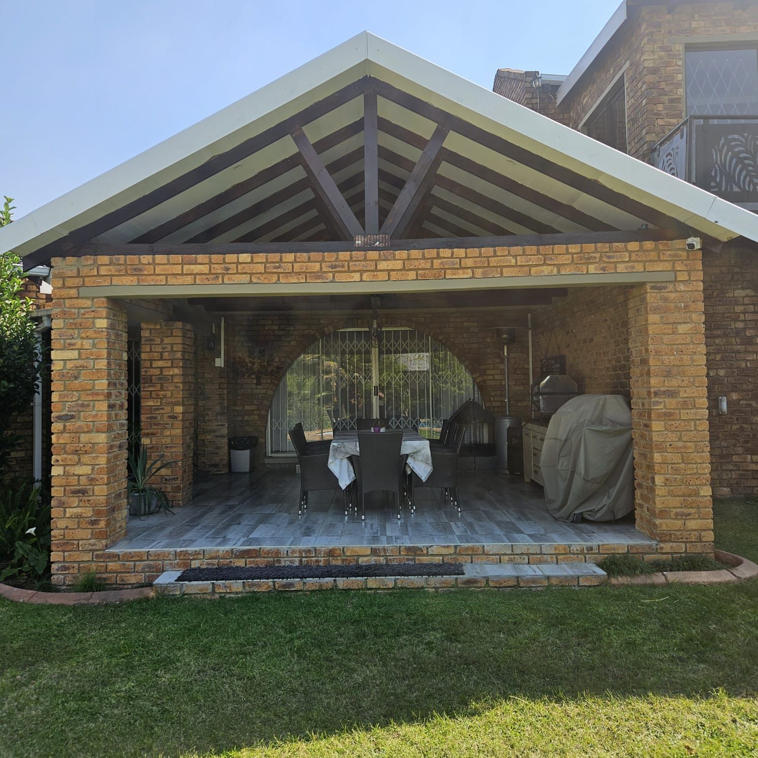 Brick patio with a gabled roof, open seating area, and grill covered at the side.