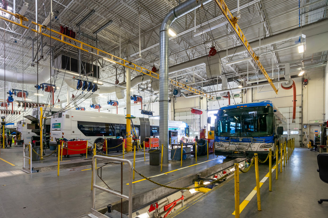 Bus in a repair bay, with tools and equipment overhead. Industrial setting with bright lights.