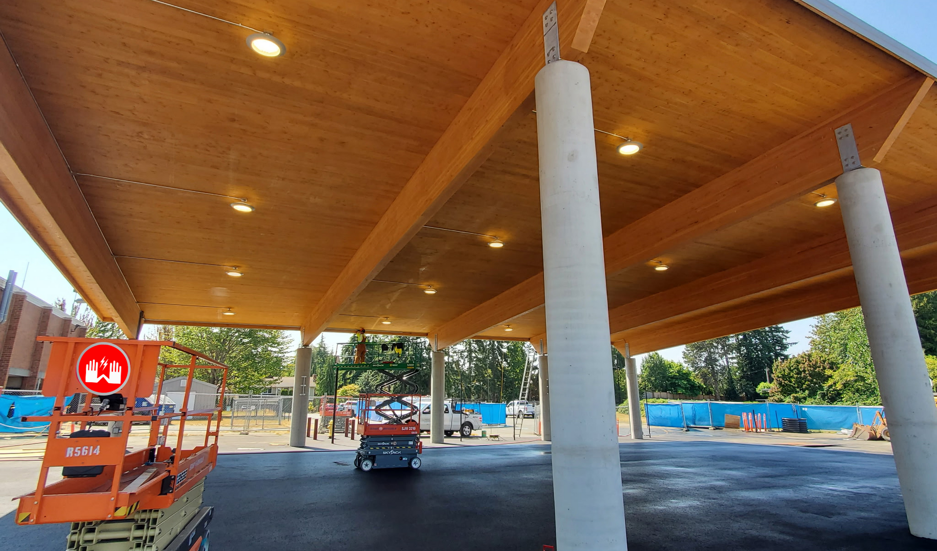 Covered parking area with wooden ceiling, concrete columns, and a scissor lift parked beneath it.