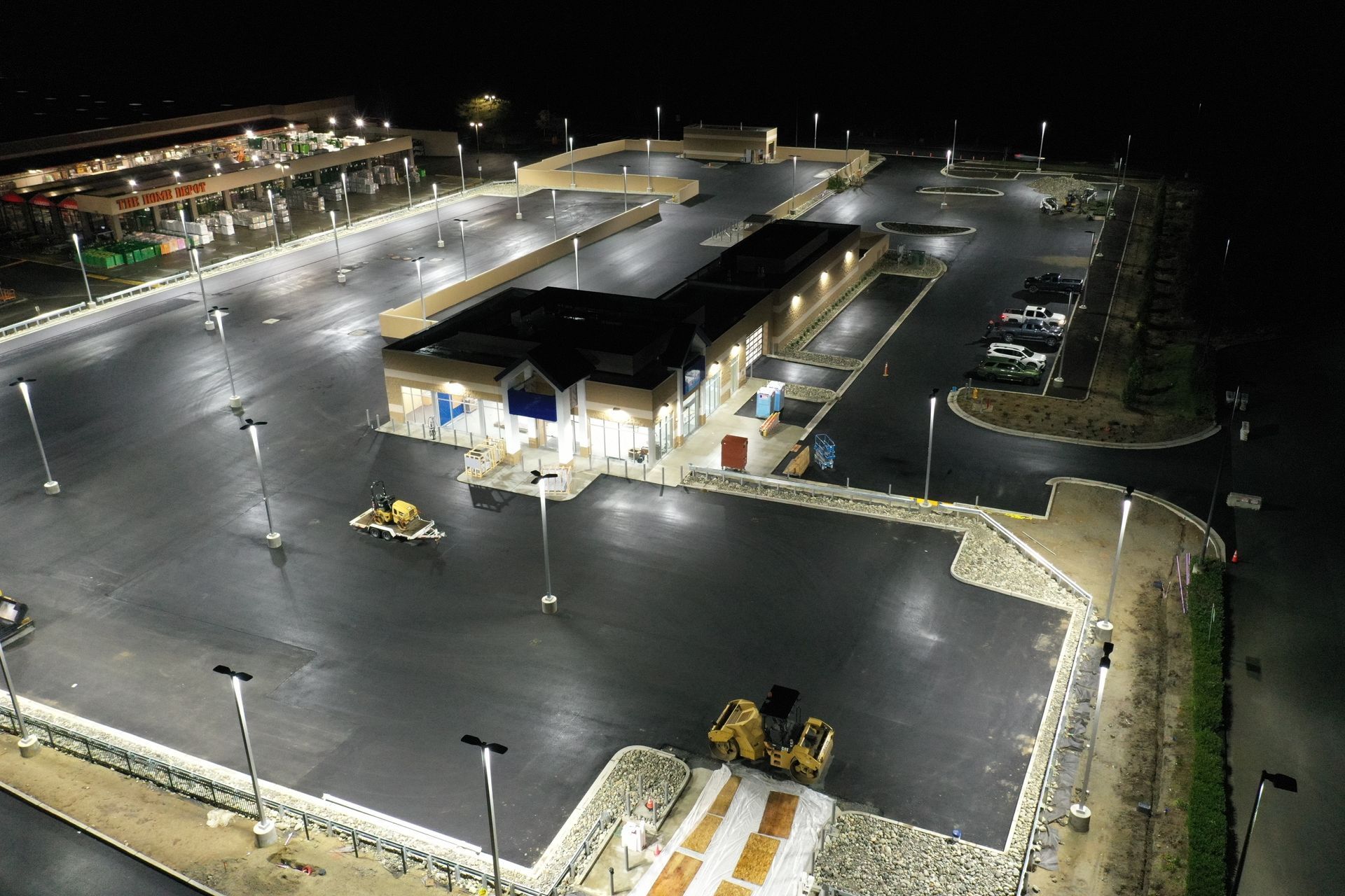 An aerial night view of a brightly lit commercial parking lot with a building and parked cars.