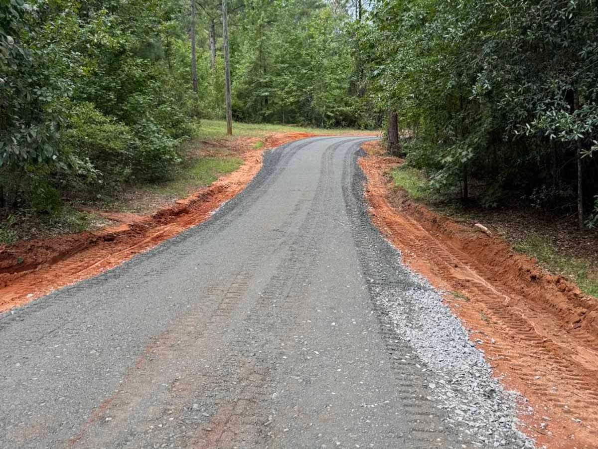 Gravel road winding through trees, with red dirt shoulders on either side.