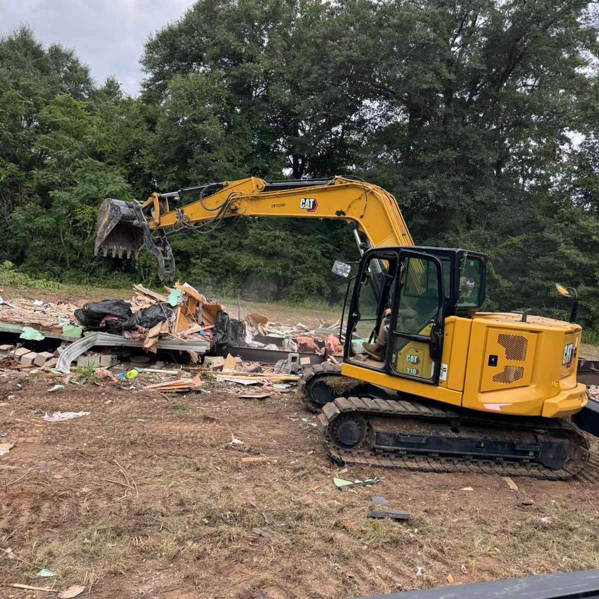 A yellow excavator demolishing a building in a grassy outdoor setting.