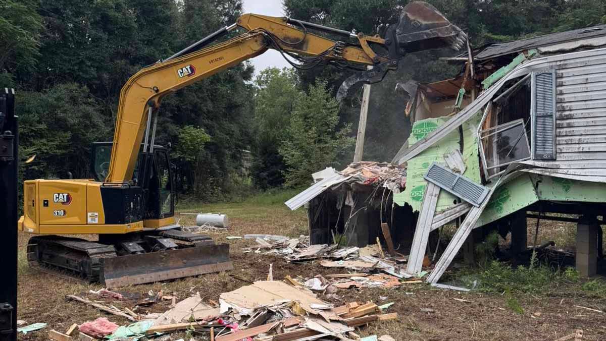 Yellow excavator demolishing a green and white house, debris on the ground. Outdoors, day.