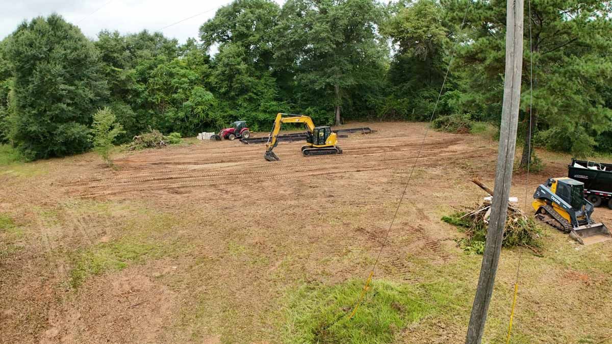 Construction site with excavator, trees cleared, and machinery on a brown, grassy area.