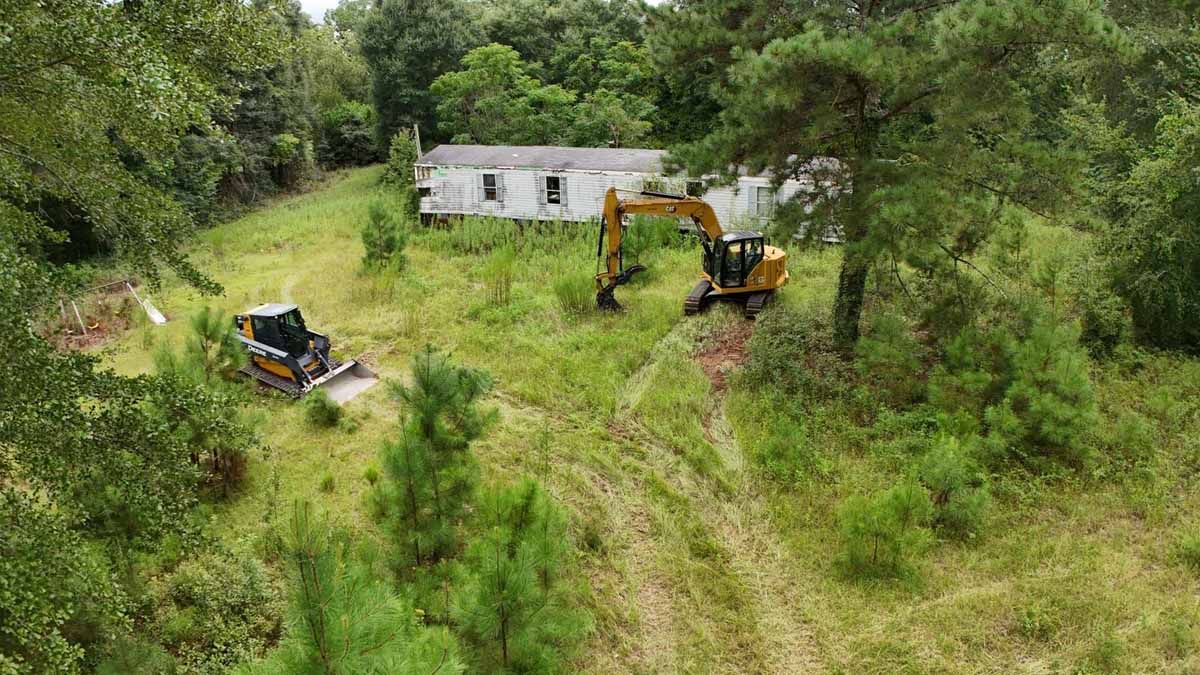 Construction equipment clearing land, a dilapidated house in the background.