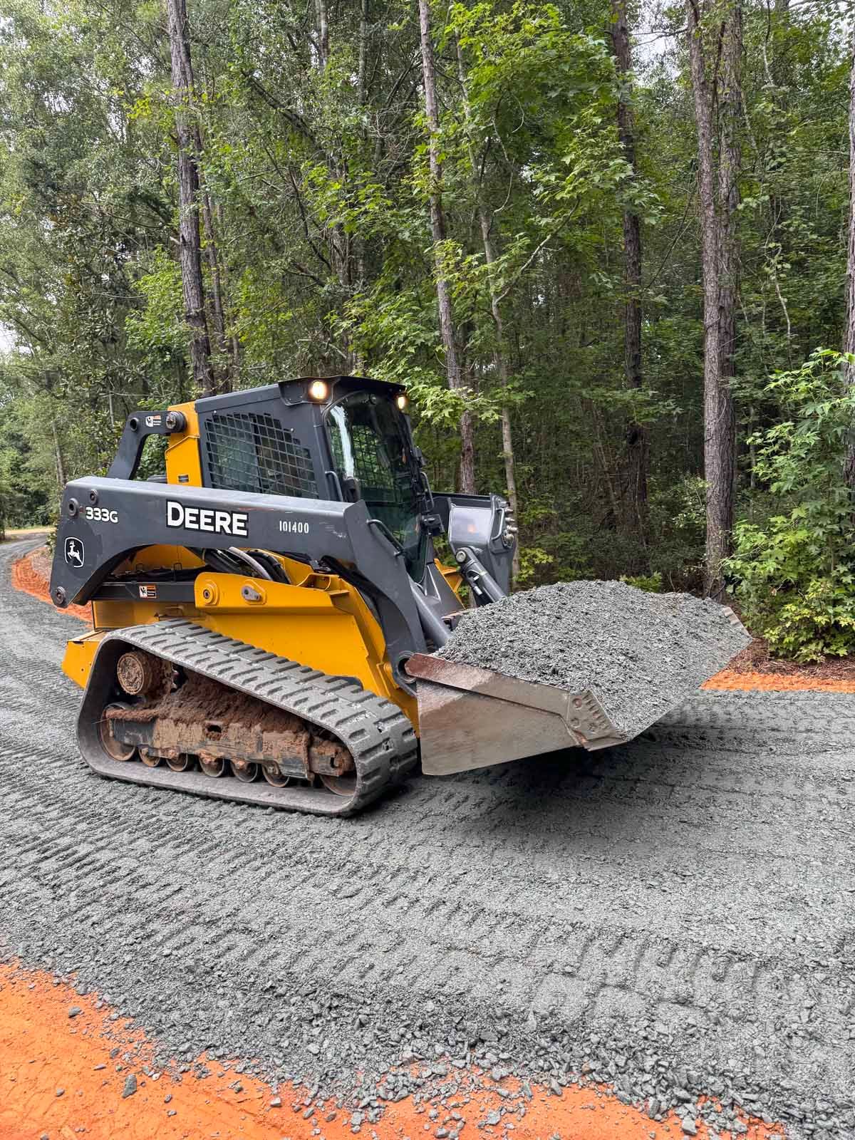 Yellow John Deere skid steer with gravel in its bucket on a gravel road in a wooded area.