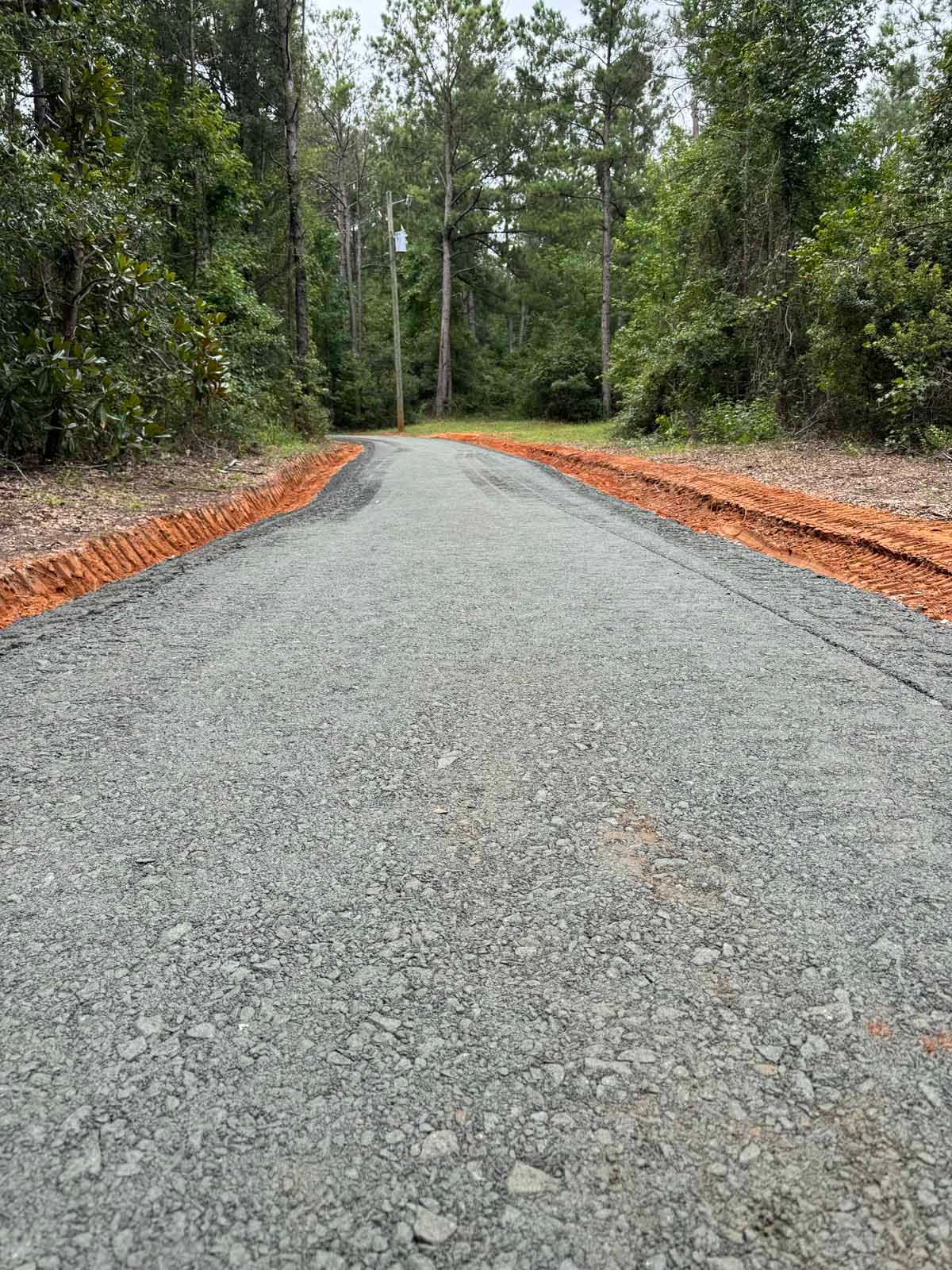 A gravel road winding through a forest, with red dirt shoulders and trees on either side.