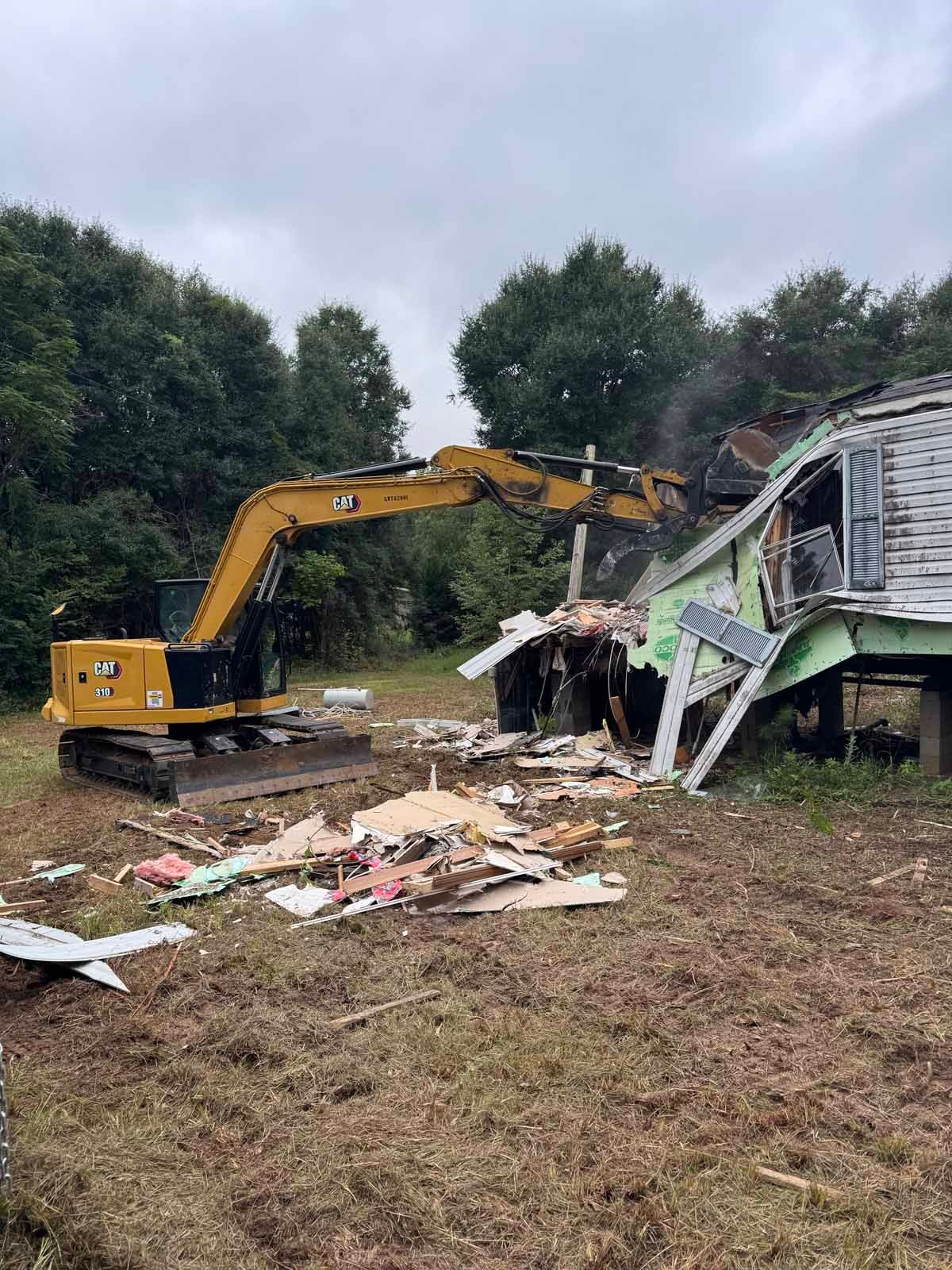 A yellow excavator demolishes a house, debris scattered on the brown grass under a cloudy sky.