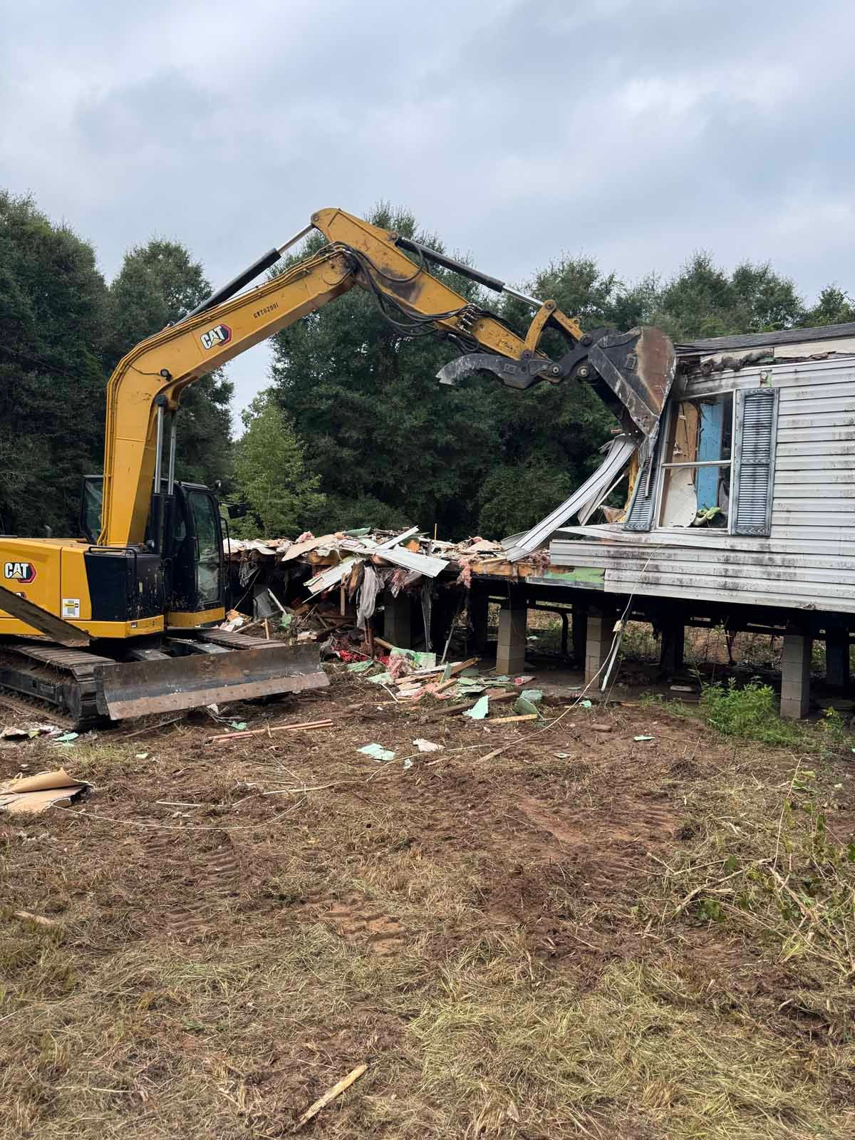 An excavator demolishes a weathered white house on stilts, with a forest in the background.