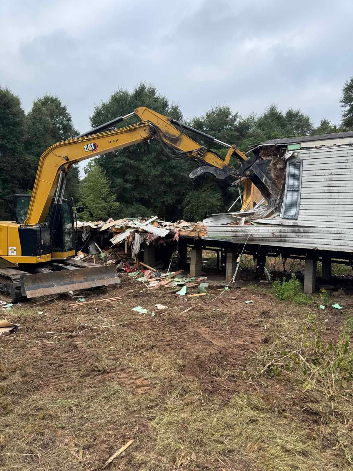 Yellow excavator demolishing a white mobile home in a grassy area. Trees in background.