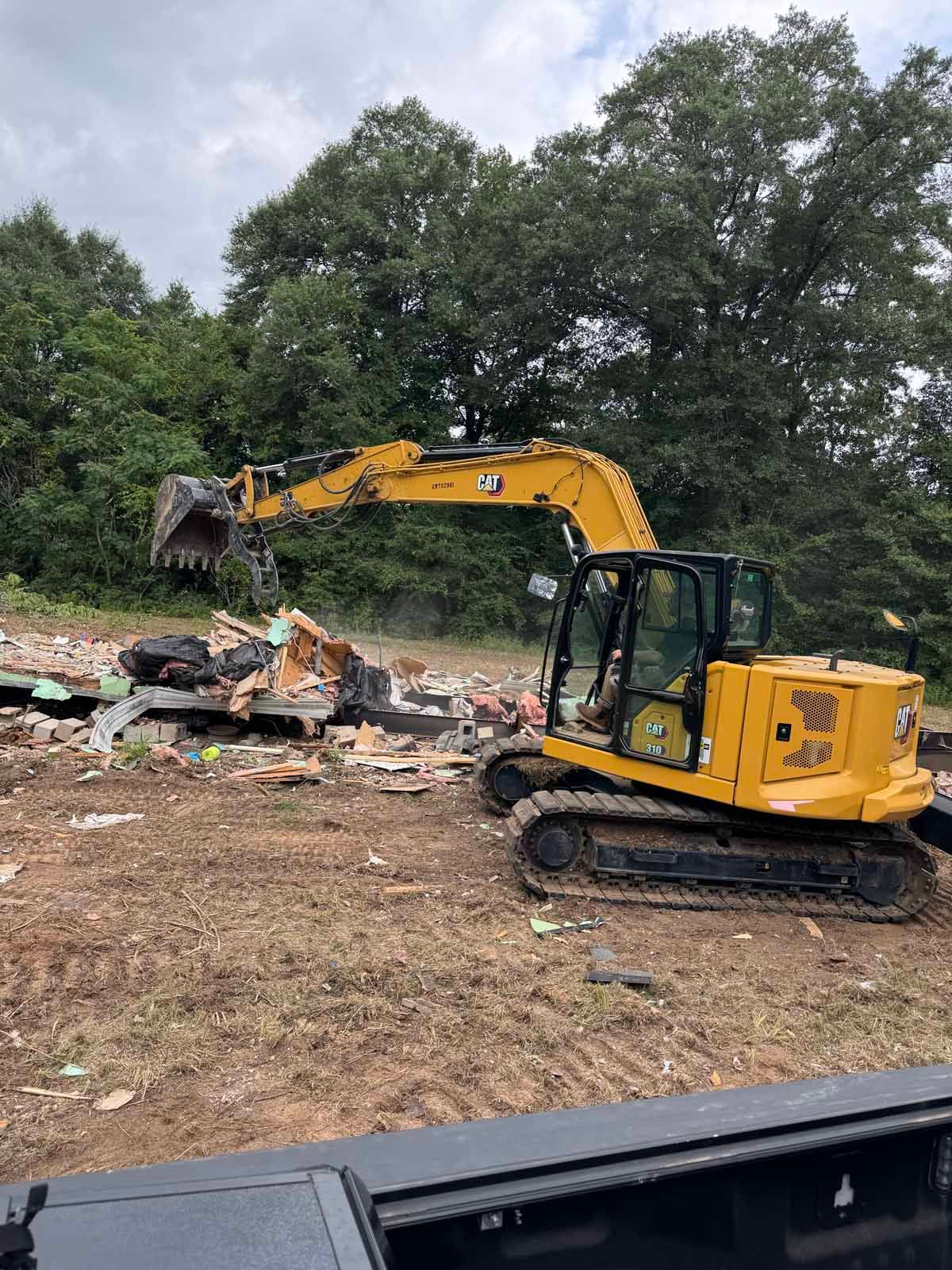 Yellow excavator demolishing a building in a field, with trees in the background, cloudy sky.