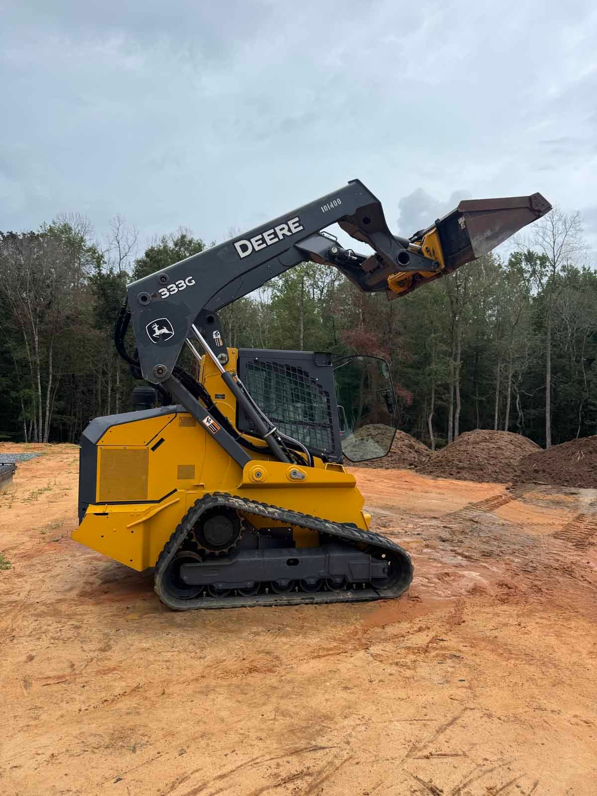 Yellow Deere skid steer with a tree shear attachment, parked on dirt, overcast sky.