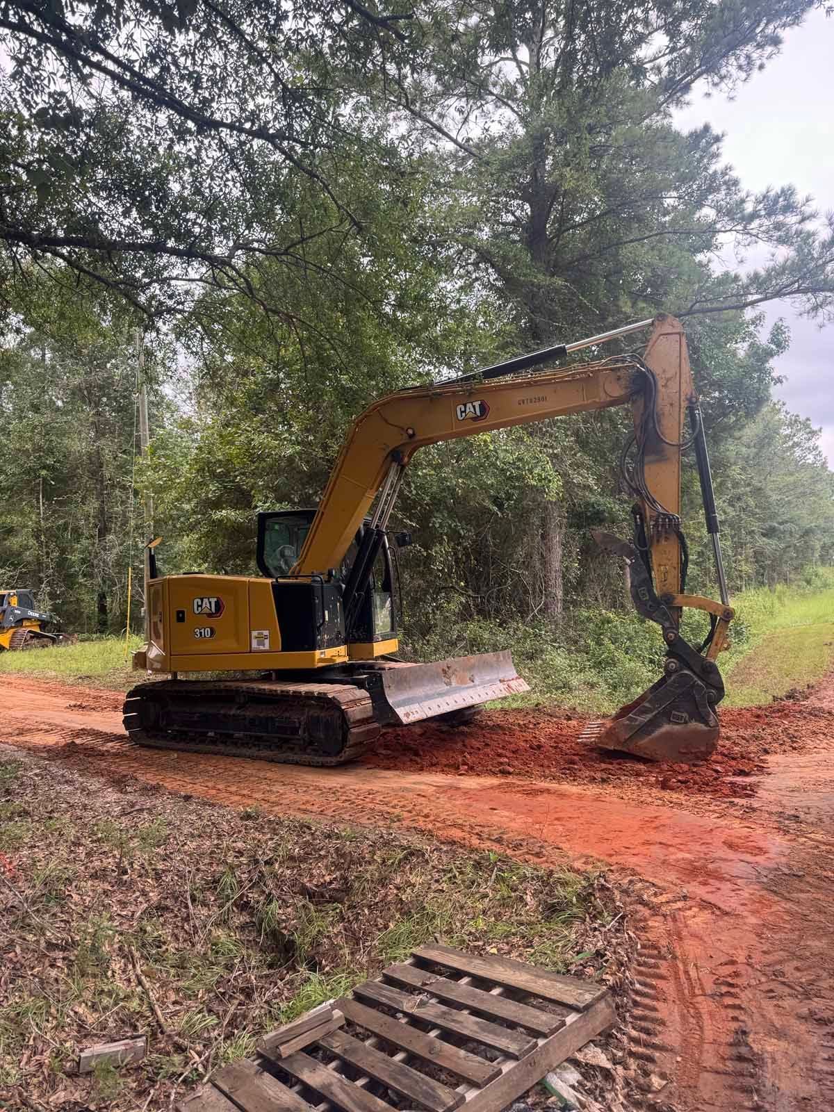 Yellow excavator on a red dirt road clearing land, trees in the background.