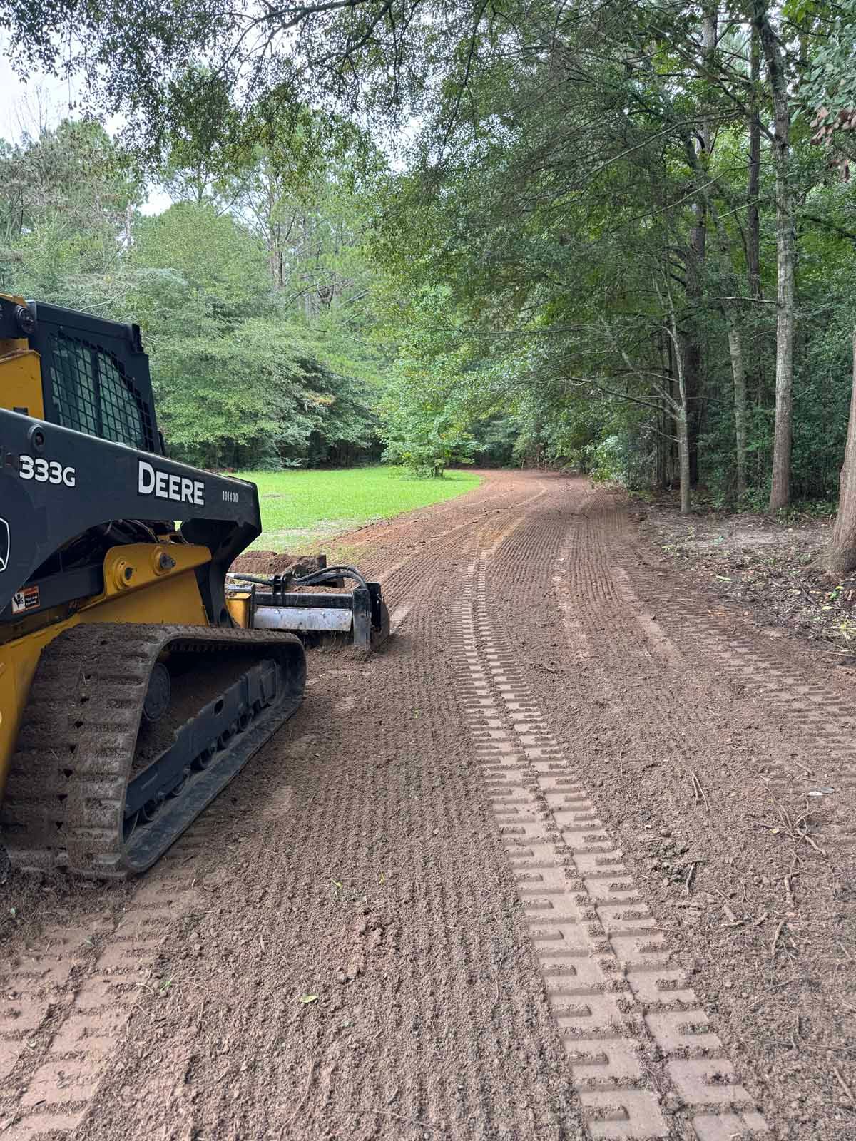 A yellow John Deere skid steer grading a muddy, winding dirt path beside a forest.