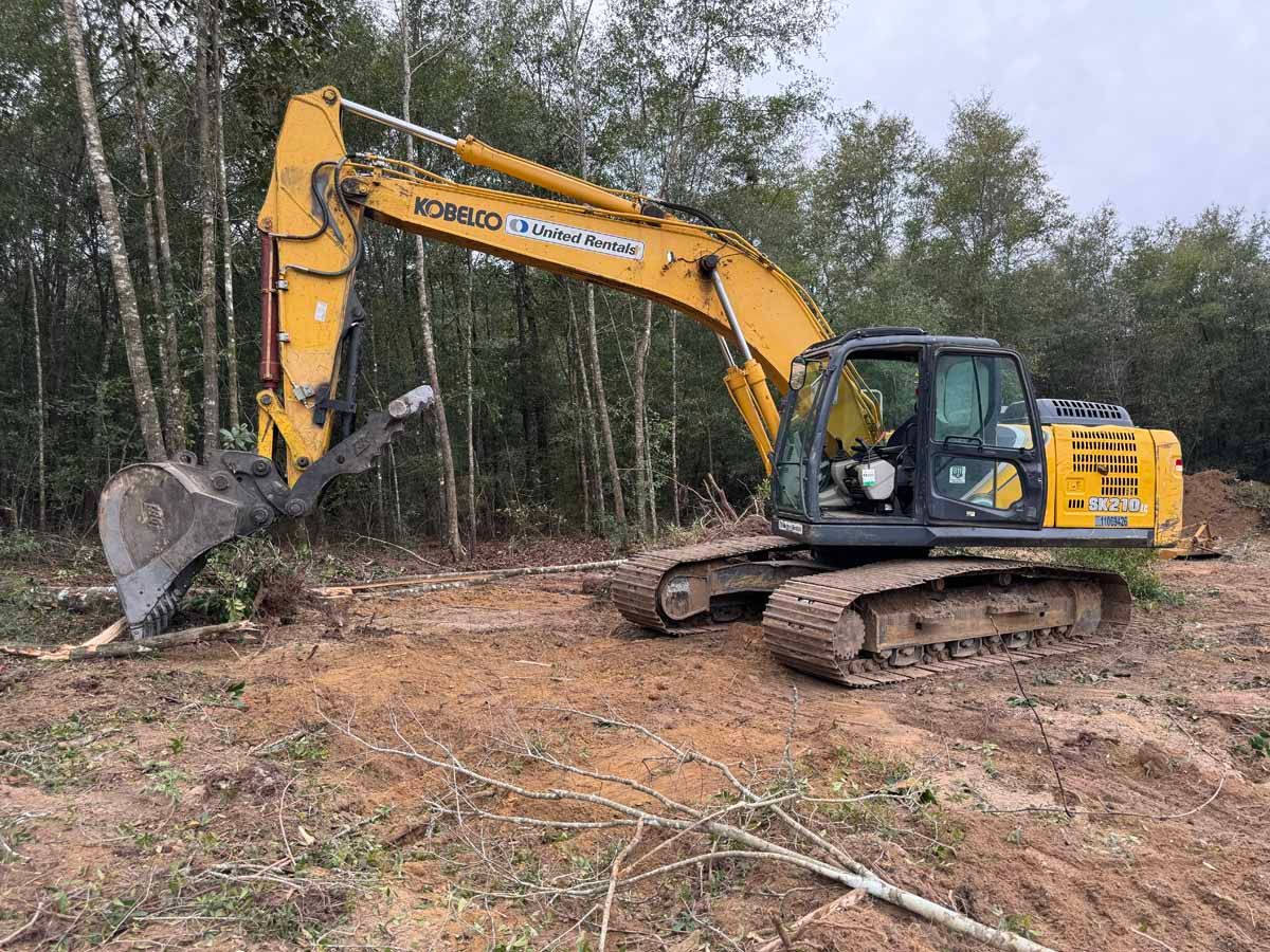 Yellow excavator on a dirt patch, clearing land near a forest.
