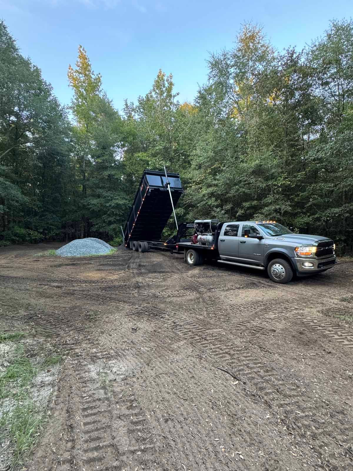 Gray truck with a raised black dump trailer unloading gravel in a wooded area.