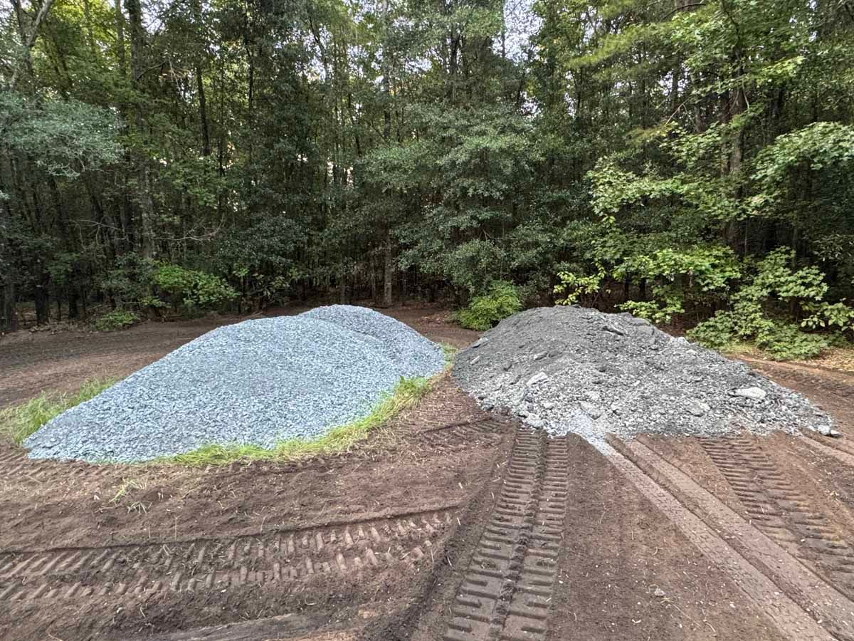 Two piles of gravel, one blue-gray and one gray, on dirt with tire tracks, set against a backdrop of green trees.