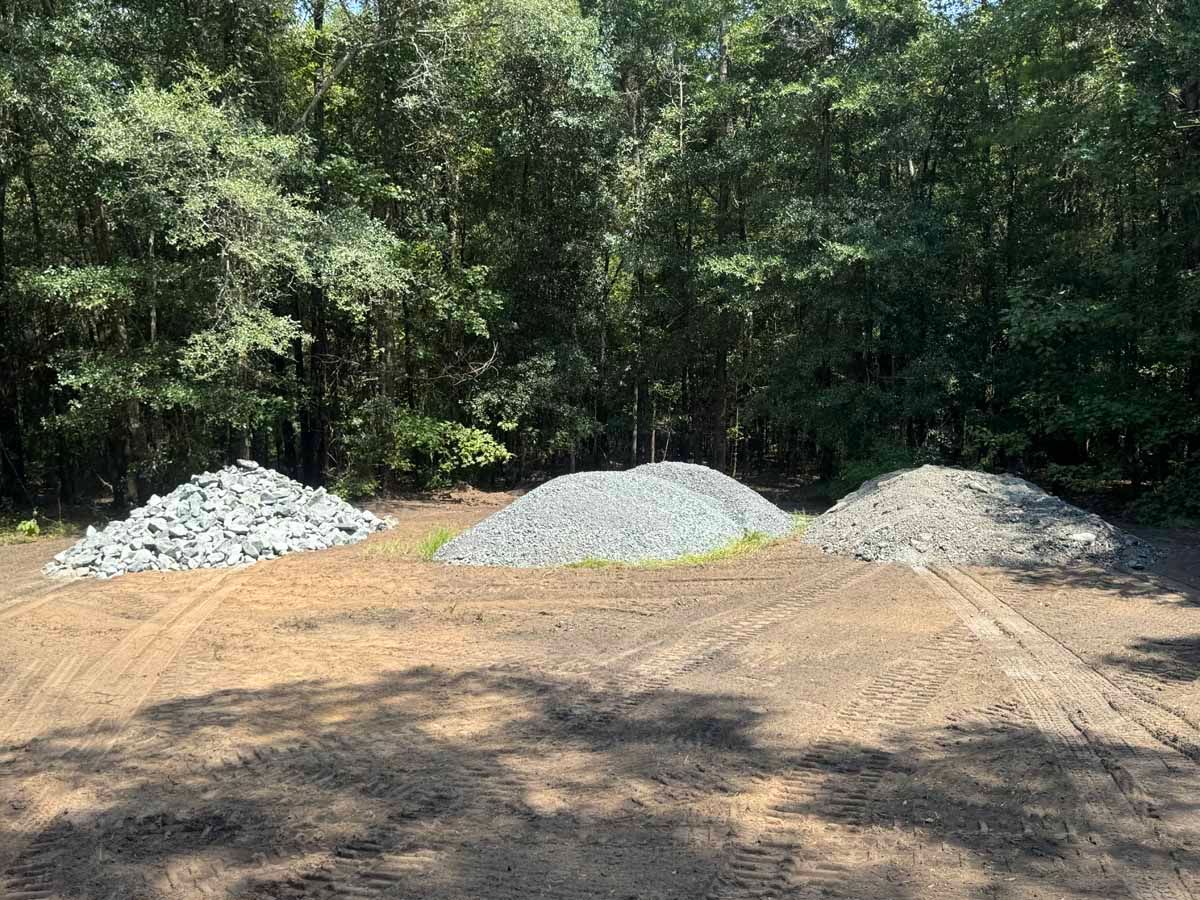 Three piles of gravel on a dirt ground, trees in the background.