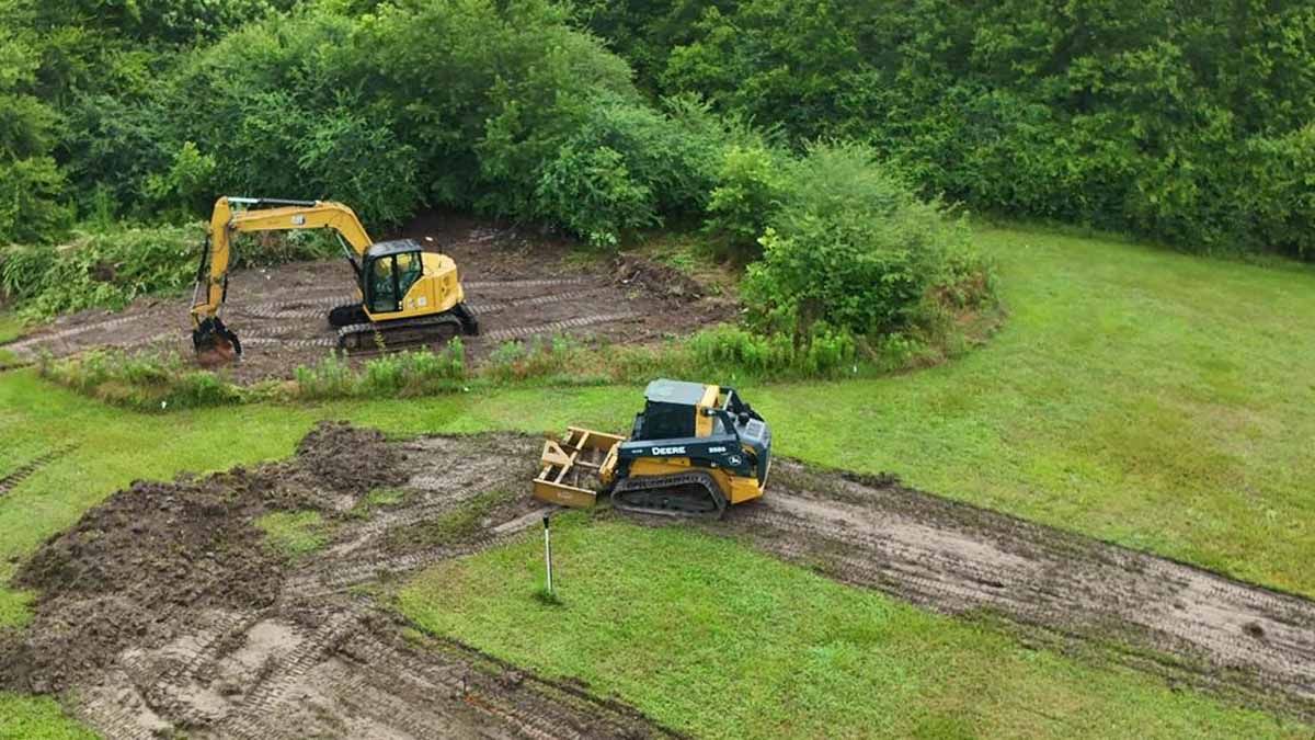 Two construction vehicles excavating mud in a grassy area near a wooded area.