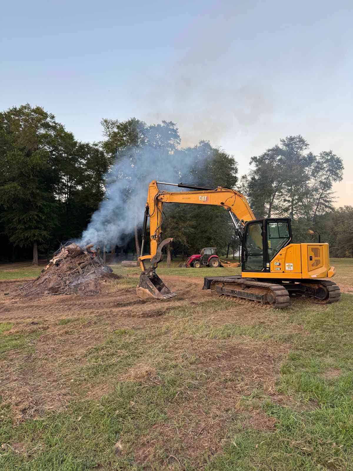 Yellow excavator moving debris near a smoky fire in a grassy field.