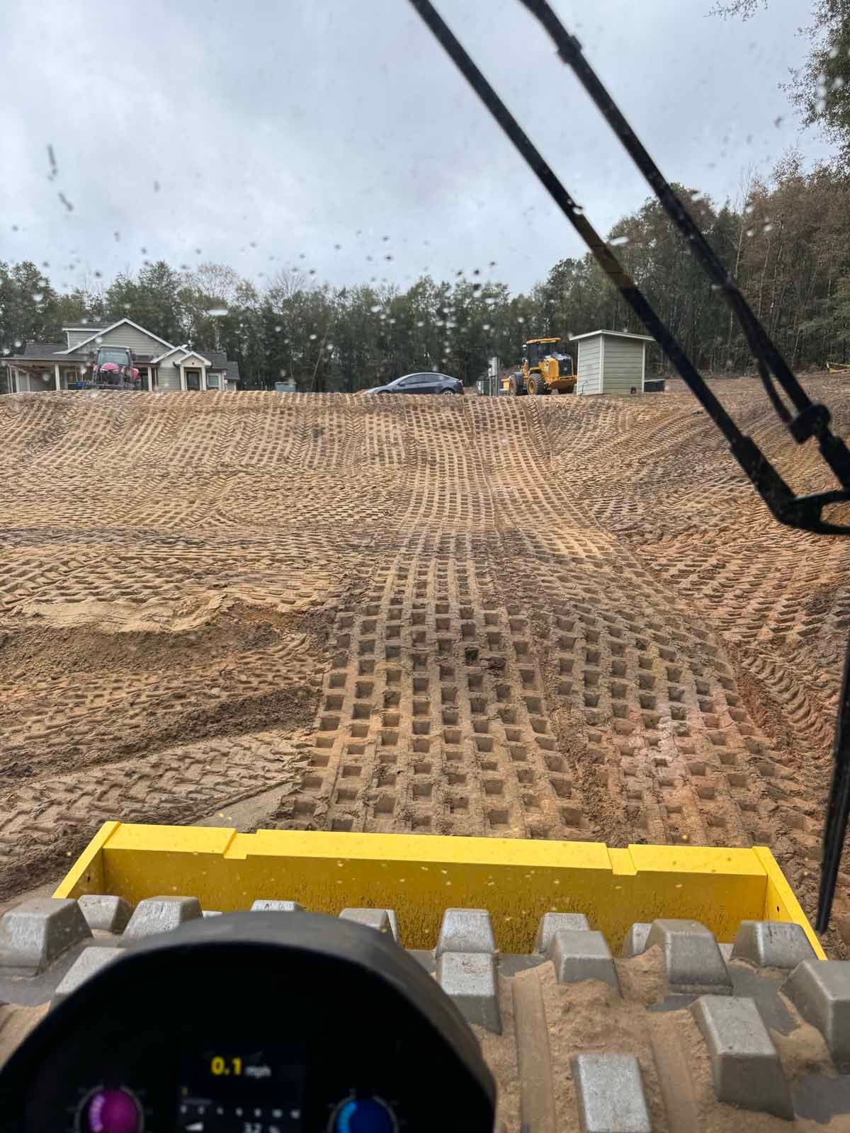 View from a vehicle of a graded construction site, with tire tracks and a building in the background.