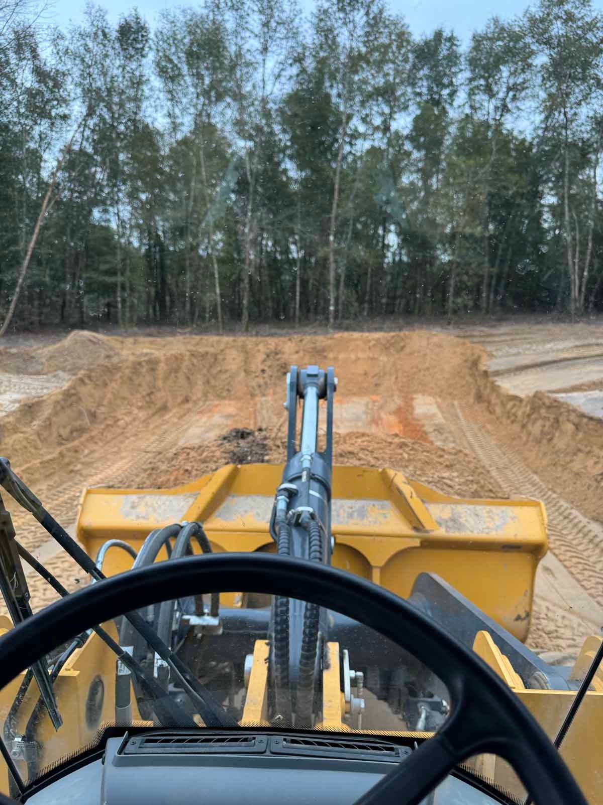 View from inside a yellow construction loader, moving dirt on a sandy lot, with a line of trees in the background.