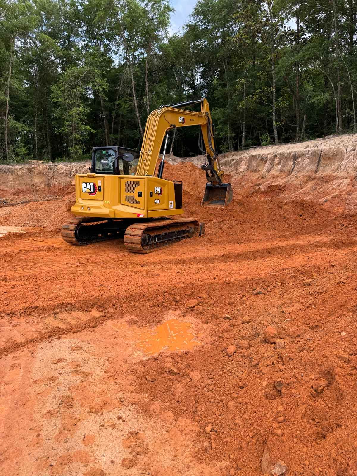 Yellow Caterpillar excavator digging in a reddish-brown dirt pit, forested background.