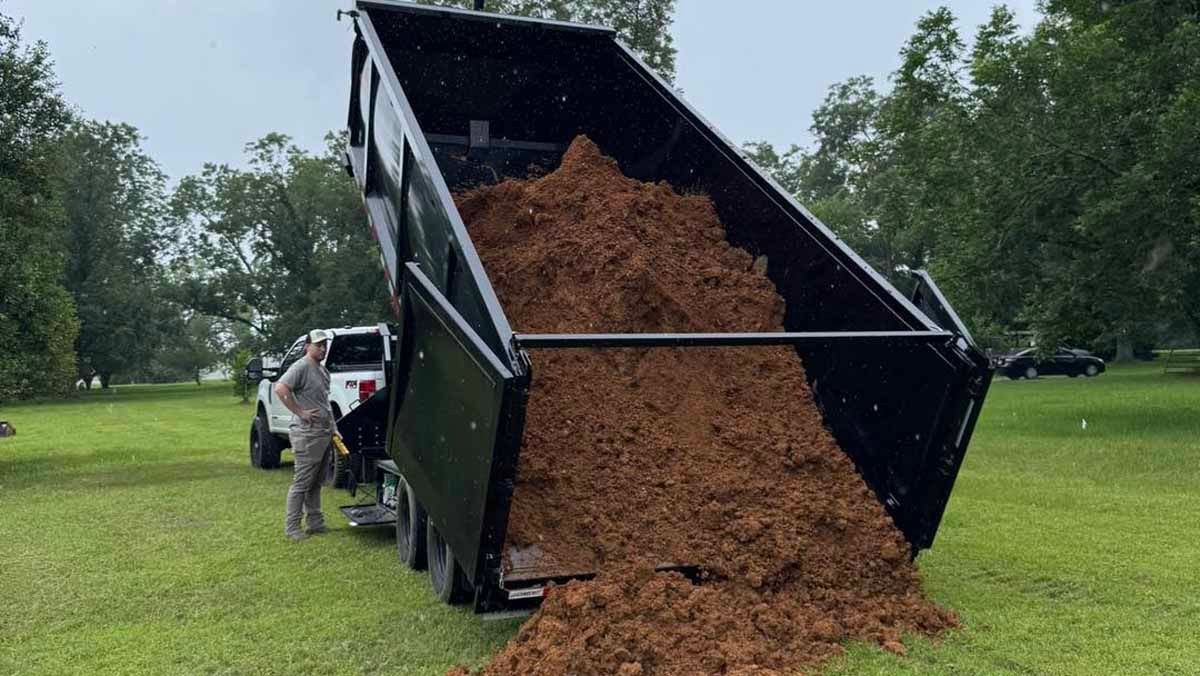 A truck trailer dumping brown mulch onto a grassy field. Man stands nearby, trees in background.