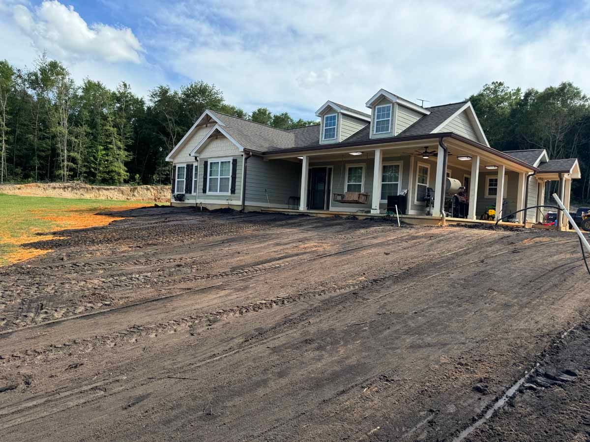 House under construction; dirt yard in front. Tan house with covered porch, dormers, and trees in background.