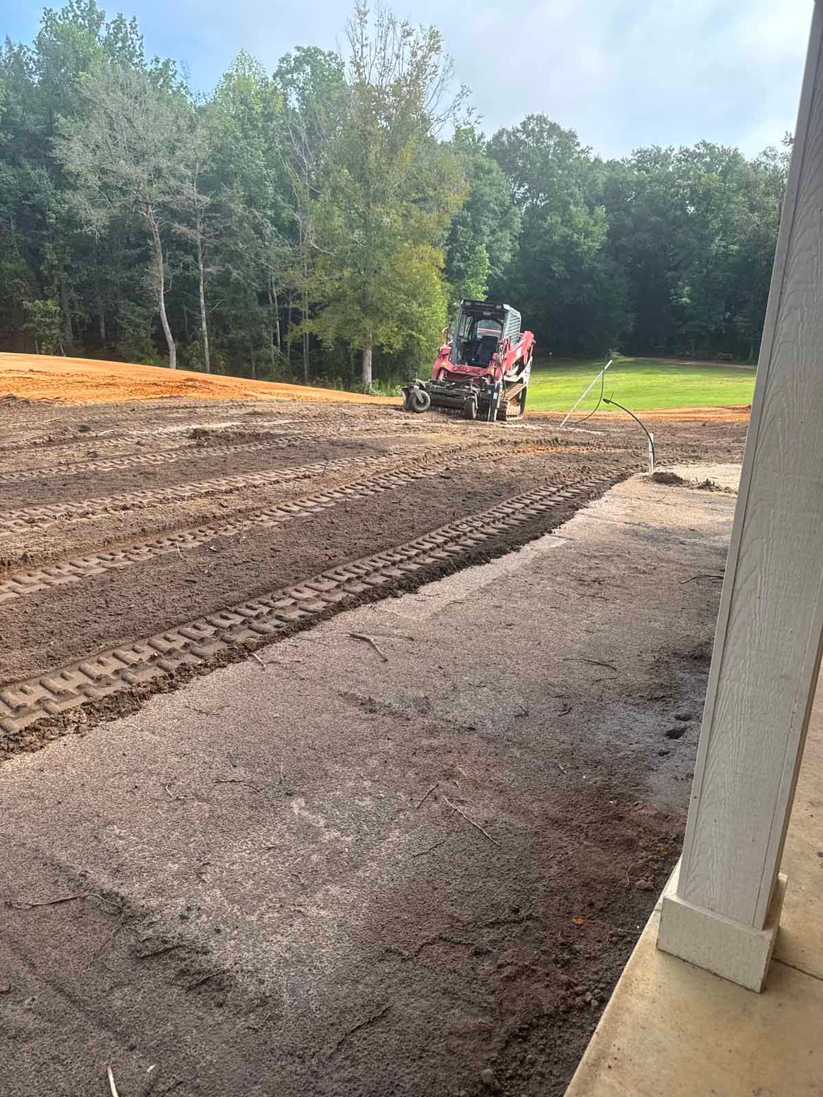 A red skid steer tractor levels dirt on a construction site. Green trees and sky in the background.