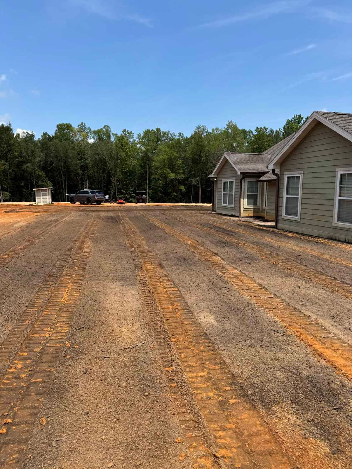 A gravel parking area with a few buildings and trees in the background under a blue sky.