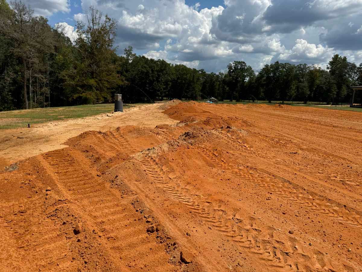Dirt field with tire tracks and a dark pipe. Trees and blue sky in the background.