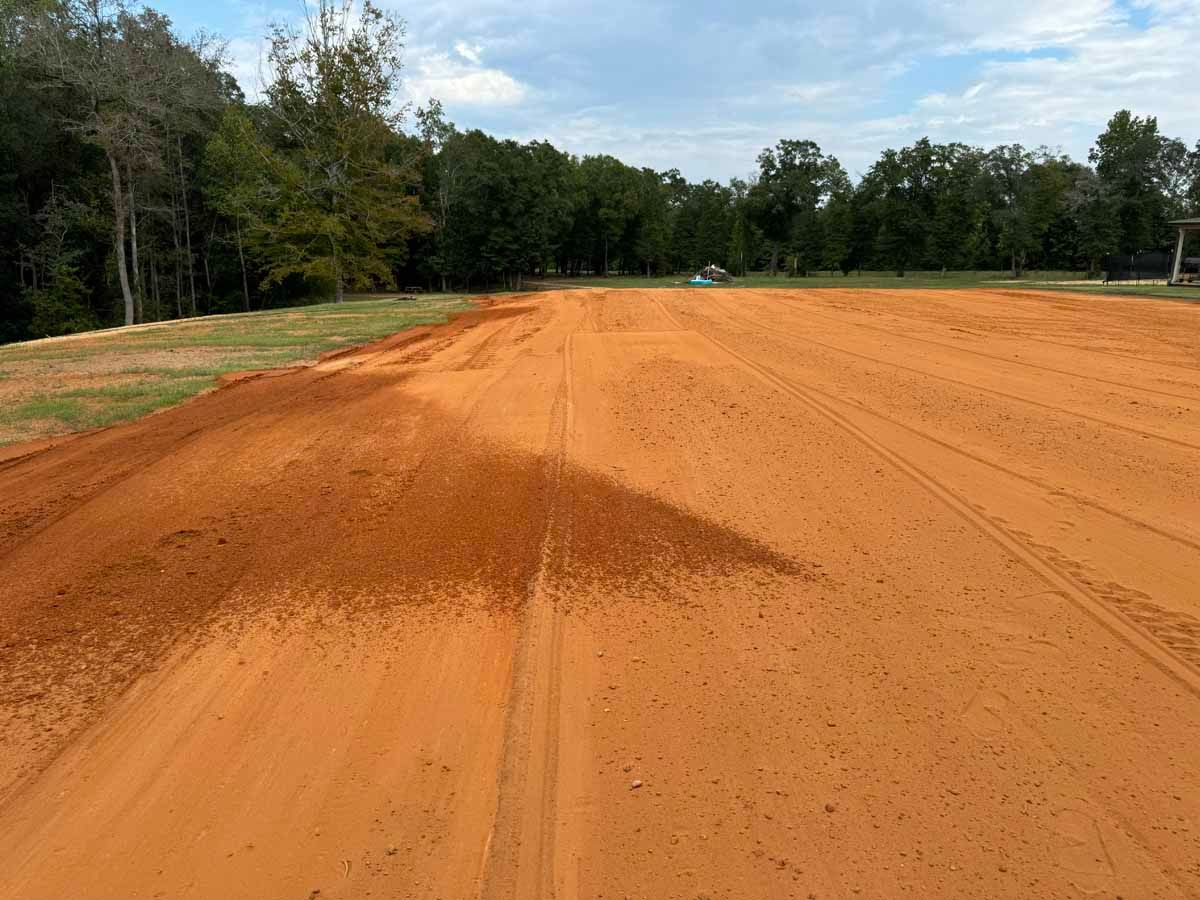 Flat, red dirt area; possibly a construction site or arena. Trees line the background.