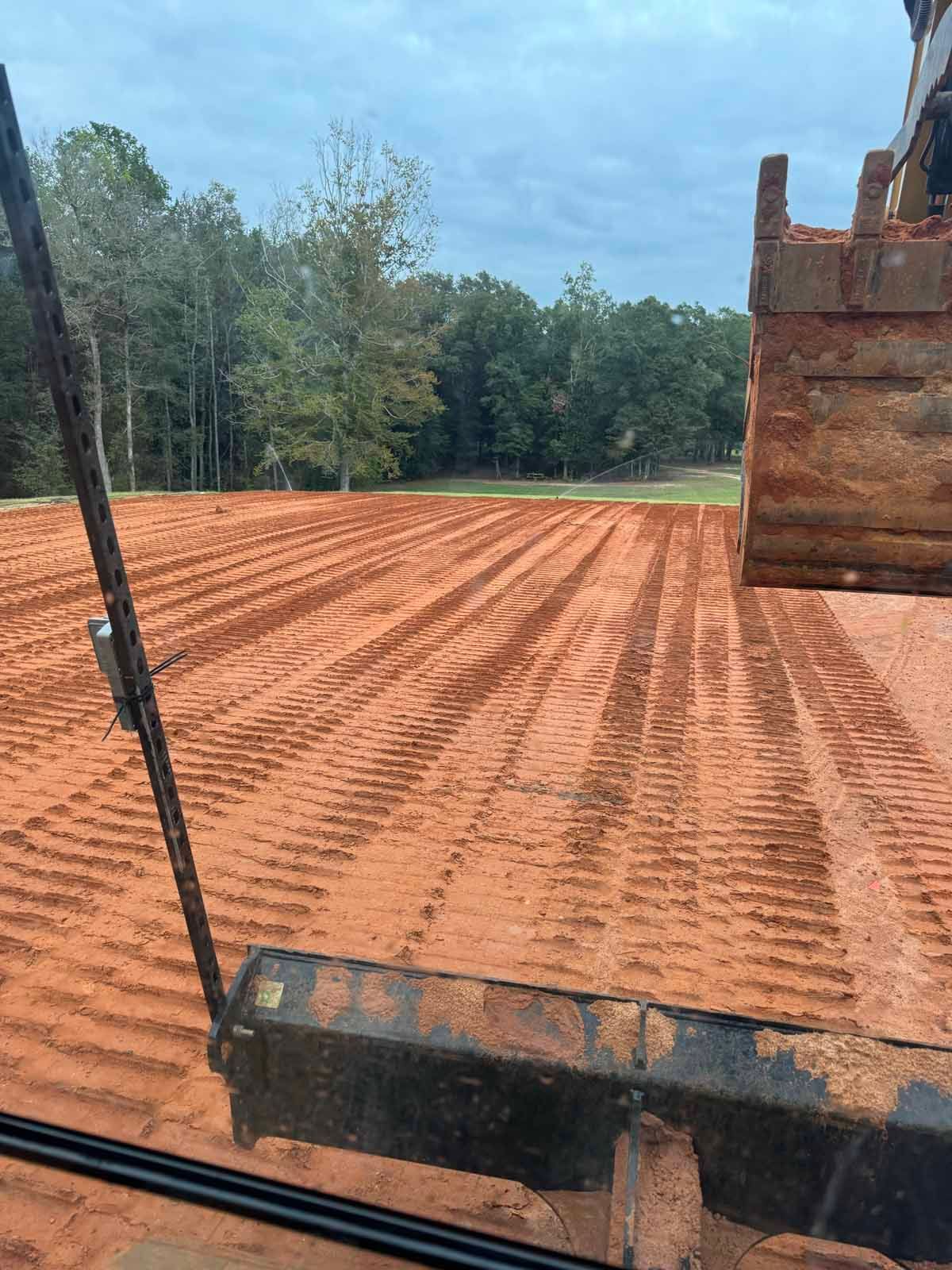 A large, flat, red dirt field, freshly graded, with a bucket in the foreground and trees in the distance.