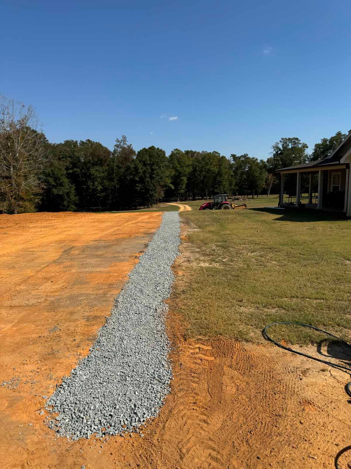 A gravel pathway bordered by dirt and grass leading to a house on a sunny day.
